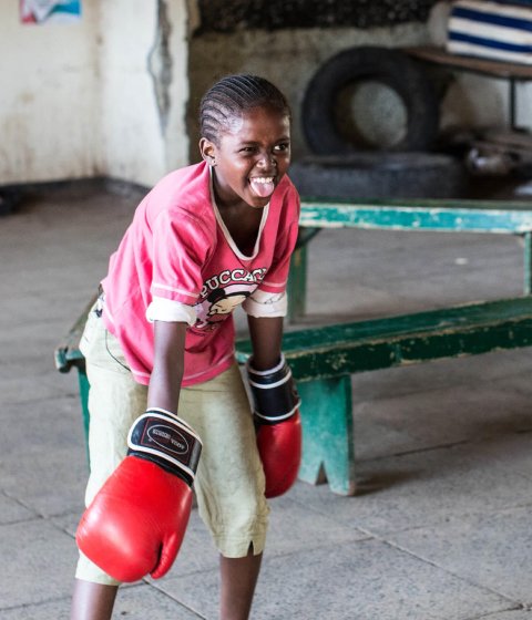 A young girl wearing boxing gloves smiles and sticks her tongue out.